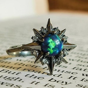 Op de afbeelding: Een zilveren ring met een stervormig ontwerp met een grote, iriserende blauwgroene opaal-edelsteen omringd door kleinere, heldere edelstenen.