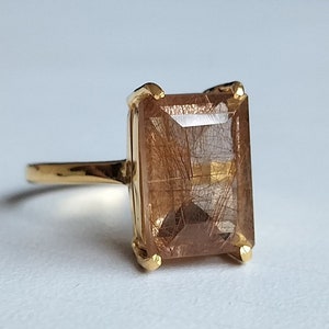 Op de afbeelding: Een gouden ring met een grote, rechthoekige, bruine edelsteen. De edelsteen heeft een unieke, getextureerde uitstraling.