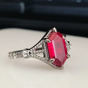 Könnte beinhalten: Ein silberner Ring mit einem großen, roten, achteckigen Edelstein. Der Ring hat ein filigranes, kompliziertes Design.