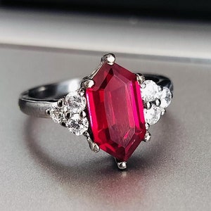 Könnte beinhalten: Ein silberner Ring mit einem großen, rechteckigen, leuchtend roten Edelstein als Mittelpunkt. Flankiert wird der rote Stein von Clustern kleinerer, klarer Edelsteine. Der Ring ist vor einem neutralen Hintergrund platziert, der sein elegantes Design hervorhebt.