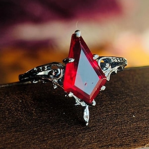 May include: A silver ring featuring a large, faceted, red gemstone in the shape of a diamond. The ring has intricate details and a gothic style.