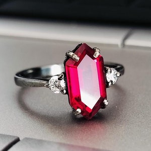 Könnte beinhalten: Ein silberner Ring mit einem großen, achteckigen, roten Edelstein und zwei kleineren, runden, weißen Edelsteinen.