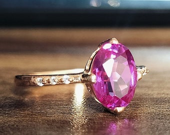 Può includere: Un anello in oro rosa con una grande pietra preziosa ovale rosa e una fascia di pietre preziose più piccole e trasparenti.