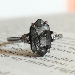 Op de afbeelding: Een zilveren ring met een zeshoekige zwarte en heldere edelsteen en twee kleinere heldere edelstenen op de band.