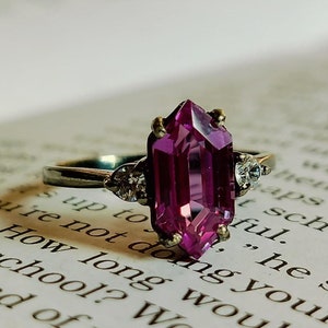 Op de afbeelding: Een zilveren ring met een grote, gefacetteerde, paarse edelsteen en twee kleinere, heldere edelstenen op de band.