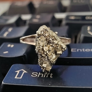 Könnte beinhalten: Ein silberner Ring mit einem großen, rauen, goldfarbenen Edelstein. Der Edelstein ist ein Cluster aus kleinen Kristallen.