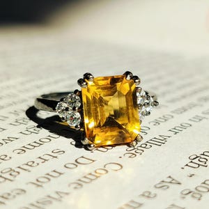 Op de afbeelding: Een zilveren ring met een grote, vierkante, gele edelsteen omringd door kleinere, heldere edelstenen.