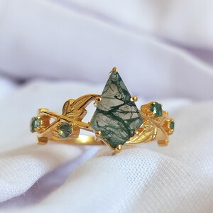 Könnte beinhalten: Ein goldener Ring mit einem grünen Moosachat-Edelstein und kleineren grünen Edelsteinen, die in einem Blattdesign eingefasst sind.