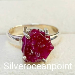 May include: A silver ring with a large, rough-cut ruby gemstone. The ruby is a deep red color and has a natural, unpolished look. The ring is set in a simple band.
