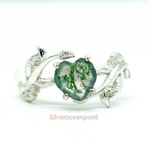 Peut inclure: Une bague en argent avec une pierre d'agate mousse verte en forme de cœur sertie dans un motif de vigne feuillue. La bague est ornée de petites pierres claires sur la vigne.