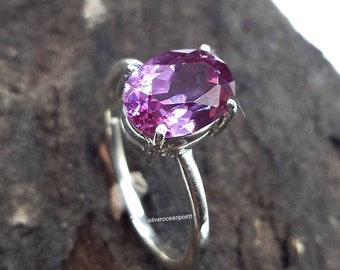 Può includere: Un anello d'argento con una grande pietra preziosa ovale di colore viola. L'anello è posto su una superficie di legno.