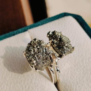 Op de afbeelding: Een zilveren ring met twee grote, gefacetteerde pyrietstenen. De stenen zijn donker goud van kleur en hebben een ruwe, natuurlijke textuur. De ring is gezet in een eenvoudige band.