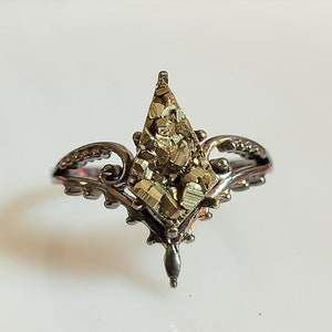 Op de afbeelding: Een zilveren ring met een grote, gefacetteerde, goudkleurige pyriet steen, gezet in een delicaat, versierd ontwerp.