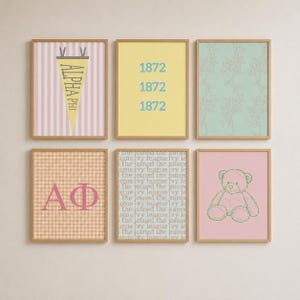 Op de afbeelding: Een set van zes ingelijste wandkunstprints. De bovenste rij bevat een roze gestreepte wimpel met de tekst ALPHA PHI, een gele print met de tekst 1872 en een mintgroen bloemenpatroon. De onderste rij toont een oranje gingham-print met de Griekse letters AΦ, een blauwe print met de herhalende tekst the joined ivy league en een roze print met een groene lijntekening van een teddybeer.
