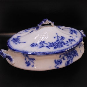 May include: A white porcelain serving dish with a blue floral pattern and a lid. The dish has a scalloped edge and two handles.