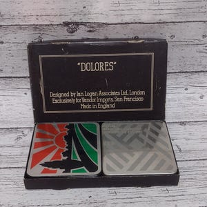 May include: A set of coasters in a black box. One coaster features a red and orange sun design with a black silhouette, and the other has a silver geometric pattern. The box reads "DOLORES" and "Made in England."