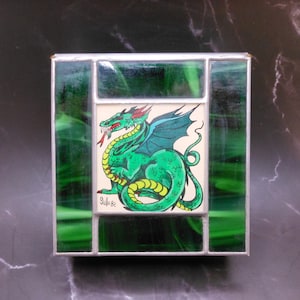 May include: A green stained glass picture frame with a green dragon illustration inside. The dragon is curled up with its tail in its mouth. The frame has a silver border and the text "Jullo 82" is in the bottom left corner.