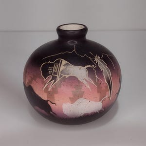 May include: A round, dark purple vase with a small opening. The vase features a carved design of a white animal, possibly a horse, with a mountain and feather motif. The background has a gradient of pink and purple.