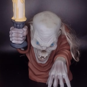 May include: A decorative figure of a pale, gaunt creature with blue eyes and long, thin hair, wearing a brown robe. The figure has one hand reaching forward and the other hand holding a tall, light-colored candle with yellow wax dripping down.