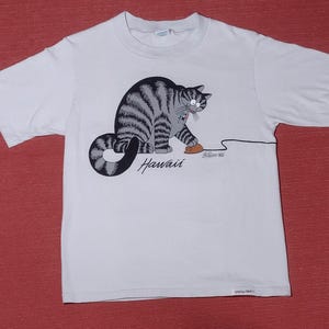 May include: A white short-sleeve t-shirt featuring a graphic of a grey tabby cat playing with a brown toy mouse. The word Hawaii is printed in black script below the cat, and a small white tag is visible at the bottom right hem of the shirt.