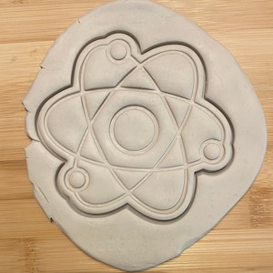May include: A white clay cookie cutter shaped like an atom. The atom has a central circle with three rings around it.