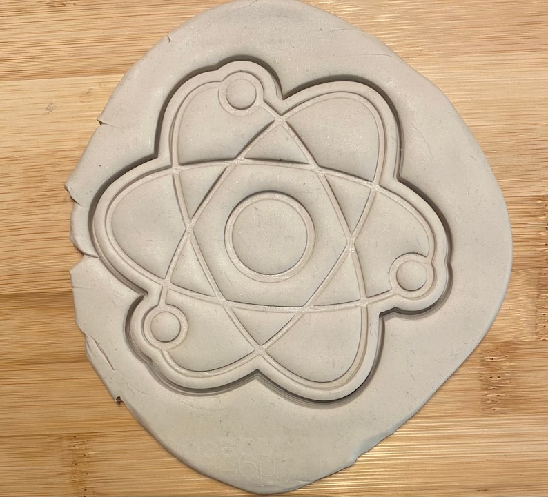 May include: A white clay cookie cutter shaped like an atom. The atom has a central circle with three rings around it.