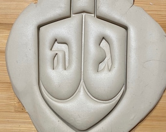 May include: A white plastic cookie cutter shaped like a dreidel, a traditional spinning top played during Hanukkah. The dreidel has the Hebrew letters "Nun", "Gimel", "Hey", and "Shin" on its sides.