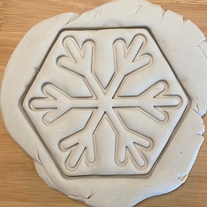 May include: A white clay imprint of a snowflake with six points. The snowflake is embossed in the clay, creating a raised design.
