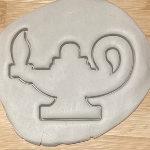 Pode incluir: Uma superfície de argila branca com um recorte de uma lâmpada de gênio. A lâmpada tem um bico curvo e uma alça.