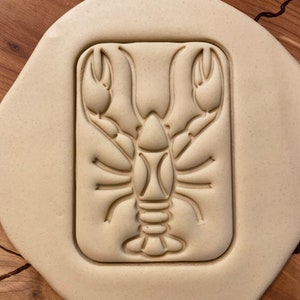 May include: A beige clay cookie cutter in the shape of a lobster. The cutter is sitting on a wooden surface.