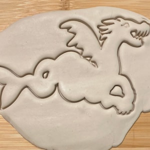 May include: A light-colored clay dragon-shaped cookie cutter. The dragon has wings, a long neck, and a fish-like tail. The cutter is pressed into a light-colored dough, creating an impression of the dragon.