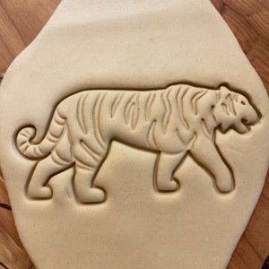 May include: A close-up of a tiger cookie cutter pressed into a light brown dough. The cutter is made of metal and has a detailed design of a tiger with stripes.