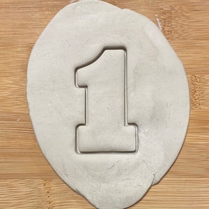 Puede incluir: Una superficie de arcilla blanca con un cortador de galletas con forma de número uno presionado en ella.