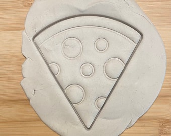May include: A white clay surface with a pizza slice-shaped cookie cutter. The cutter has a design of pepperoni circles.
