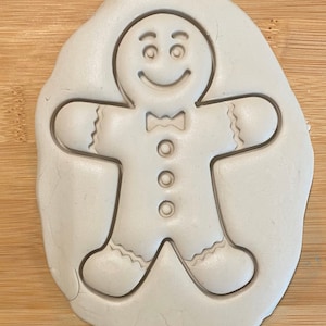 May include: A white gingerbread man cookie cutter with a smiling face, bow tie, and four buttons. The cutter is on a wooden surface.