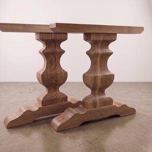 May include: Two matching brown wooden table bases featuring a central turned pedestal column with a curved silhouette, supported by a wide, contoured base and topped with a flat, rectangular horizontal support beam for a tabletop.