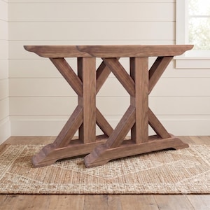 May include: A pair of brown wooden table legs featuring an X-shaped support structure. Each leg assembly consists of a horizontal top beam, a vertical central post, and two diagonal braces that connect to a wide, flat base with curved ends.