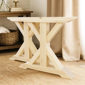 May include: Two unfinished wooden table bases. The bases are made of light-colored wood and feature an X-shaped design for support. The table bases are designed to be used for a dining table or a similar piece of furniture.
