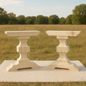 May include: Two unfinished wooden table bases with a light natural wood color. Each base has a rectangular top, a decorative central column, and a curved base. The tables are displayed on a light-colored surface in a grassy field.