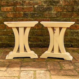 May include: Two light-colored wooden table legs featuring a central vertical support flanked by two curved side supports. Each leg has a wide, flat top piece and a sturdy, flared base with beveled edges, constructed from unfinished natural wood.
