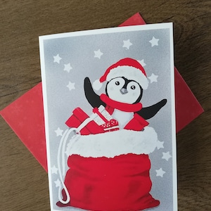 May include: A Christmas card featuring a penguin wearing a Santa hat and holding a red gift bag with white fur trim. The penguin is surrounded by white stars on a grey background.