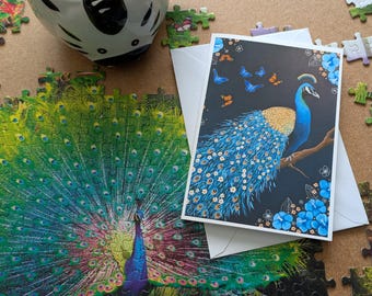 以下が含まれることがあります： 孔雀のイラストが描かれたグリーティングカードと、部分的に組み立てられた孔雀のパズル。カードには、金色の胸を持つ青い孔雀が枝に止まり、蝶と青い花が描かれています。部分的に組み立てられたパズルも表示されています。