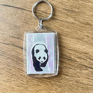 Puede incluir: Un llavero de plástico transparente con una imagen de un panda blanco y negro en su interior. El panda camina sobre un fondo de bambú rosa y blanco.