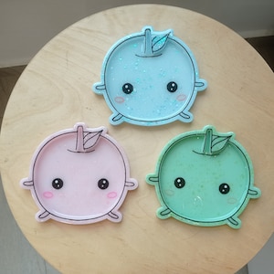 以下が含まれることがあります： かわいい顔をした樹脂製のリンゴ型のコースター3つ。コースターは青、ピンク、緑で、樹脂の中にキラキラ光るものが埋め込まれています。