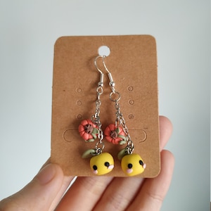 Könnte beinhalten: Ein Paar Hängeohrringe mit gelben und rosafarbenen Früchten aus Polymer Clay mit grünen Blättern und roten Blüten. Die Ohrringe sind an silbernen Ketten befestigt.