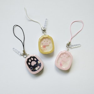 May include: Three colorful phone charms with paw print designs. One is black with a black cord, one is yellow with a white cord, and one is pink with a pink cord. Each charm has a clear, rectangular case with a paw print inside.
