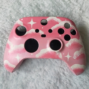 Peut inclure: Une manette Xbox personnalisée peinte en rose et blanc avec un motif de nuages et d'étoiles.