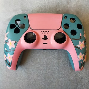 May include: A custom-painted video game controller in pink and teal. The controller has a pink centre and teal side panels. Cherry blossom branches with pink flowers are painted on the teal panels. The controller has black buttons and a black touchpad.