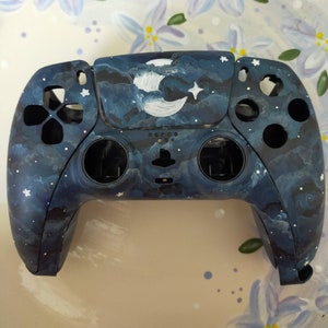 May include: A custom-painted video game controller with a dark blue, starry night sky design. The controller features a white crescent moon and stars painted on the top.
