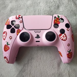 May include: A pink PlayStation 5 controller with a strawberry and flower design. The controller has black buttons and a black PlayStation logo.